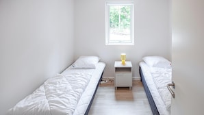 Ein kleines, sauberes Zimmer mit zwei Einzelbetten, einem Nachttisch mit Lampe und einem Fenster, das für natürliches Licht sorgt.