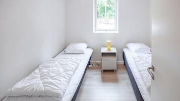 Une petite chambre propre, avec deux lits simples, une table de chevet avec une lampe et une fenêtre laissant entrer la lumière naturelle.