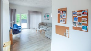 Ein modernes Wohnzimmer mit einer Couch, einem blauen Sessel, einem Couchtisch und einem großen Fenster mit Vorhängen. An den Wänden befinden sich Korkwände mit Bildern und Notizen.