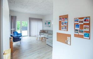 Una sala moderna con un sofá, un sillón azul, una mesita de centro y un ventanal con cortinas. Se ven tapices con fotos y notas en las paredes.