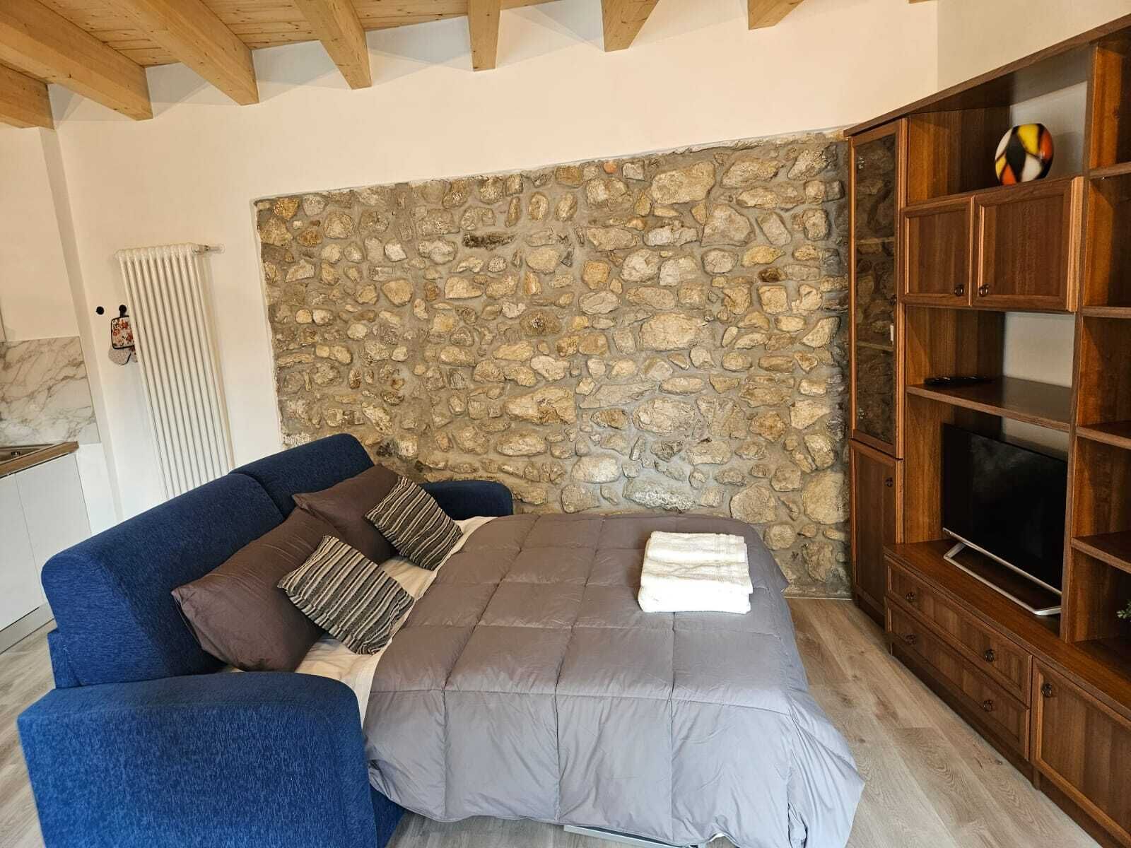 Ett sovrum med en vägg av sten, en säng med ett grått täcke, en blå soffa och ett träskåp med en tv.