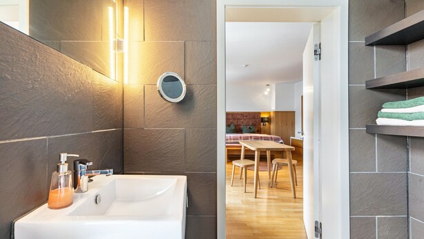 Une salle de bain moderne avec un grand lavabo, un miroir et un miroir rond accroché au mur. La pièce adjacente contient une table et des chaises en bois.