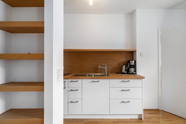 Cocina compacta con encimera de madera, armarios blancos y pileta de acero inoxidable, además de una cafetera.
