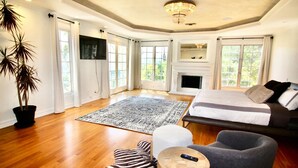 A spacious bedroom with a large bed, a fireplace, a TV, and a view of greenery through large windows. - Spacious 6-Bedroom Estate in Fabulous Los Angeles with Tennis ,Pool & Spa (Los Angeles)