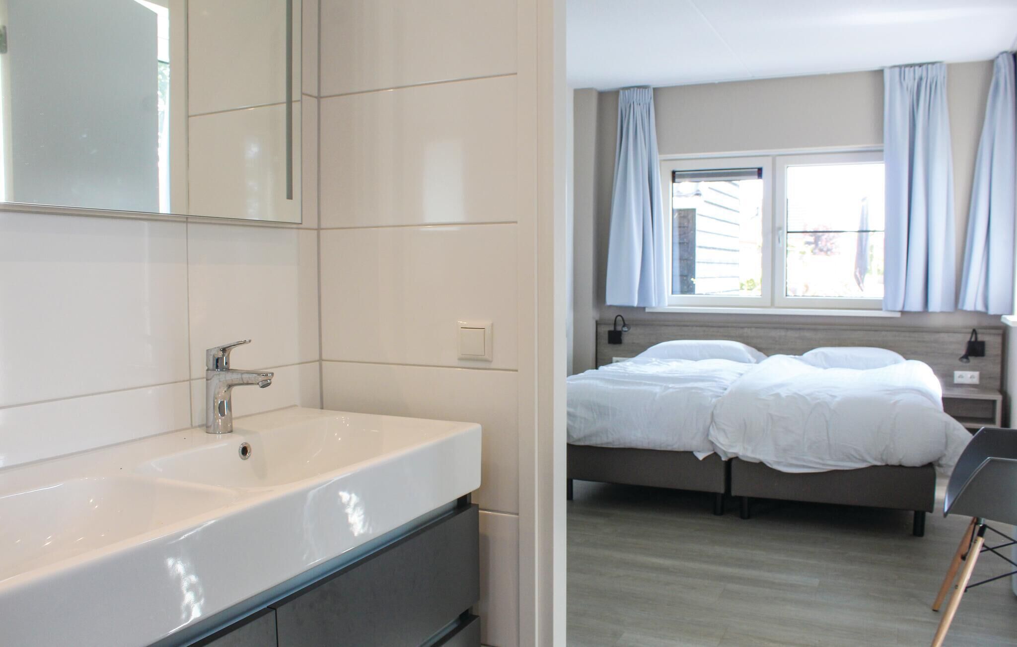 Ein modernes Badezimmer mit einem großen, weißen Waschbecken und Spiegel, einer separaten Duschkabine sowie ein Schlafzimmer mit einem Doppelbett und einem Fenster mit Vorhängen.