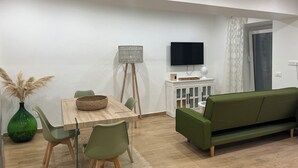 A modern living room with a green sofa, a wooden dining table with chairs, a floor lamp, and a TV mounted on the wall.