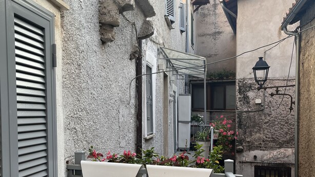 A small outdoor balcony with a table and chairs, potted plants, and a view of adjacent buildings.