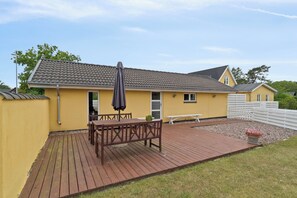 A yellow building with a tiled roof, a wooden deck, a table with chairs, and a bench. - Apartment in Snogebæk near the beach (Nexø)