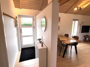 Ein Zimmer mit Holzboden, einer weißen Tür mit schwarzer Fußmatte, einem Esstisch mit Stühlen, einem Fernseher auf einem Schrank und einem Spiegel an der Wand.