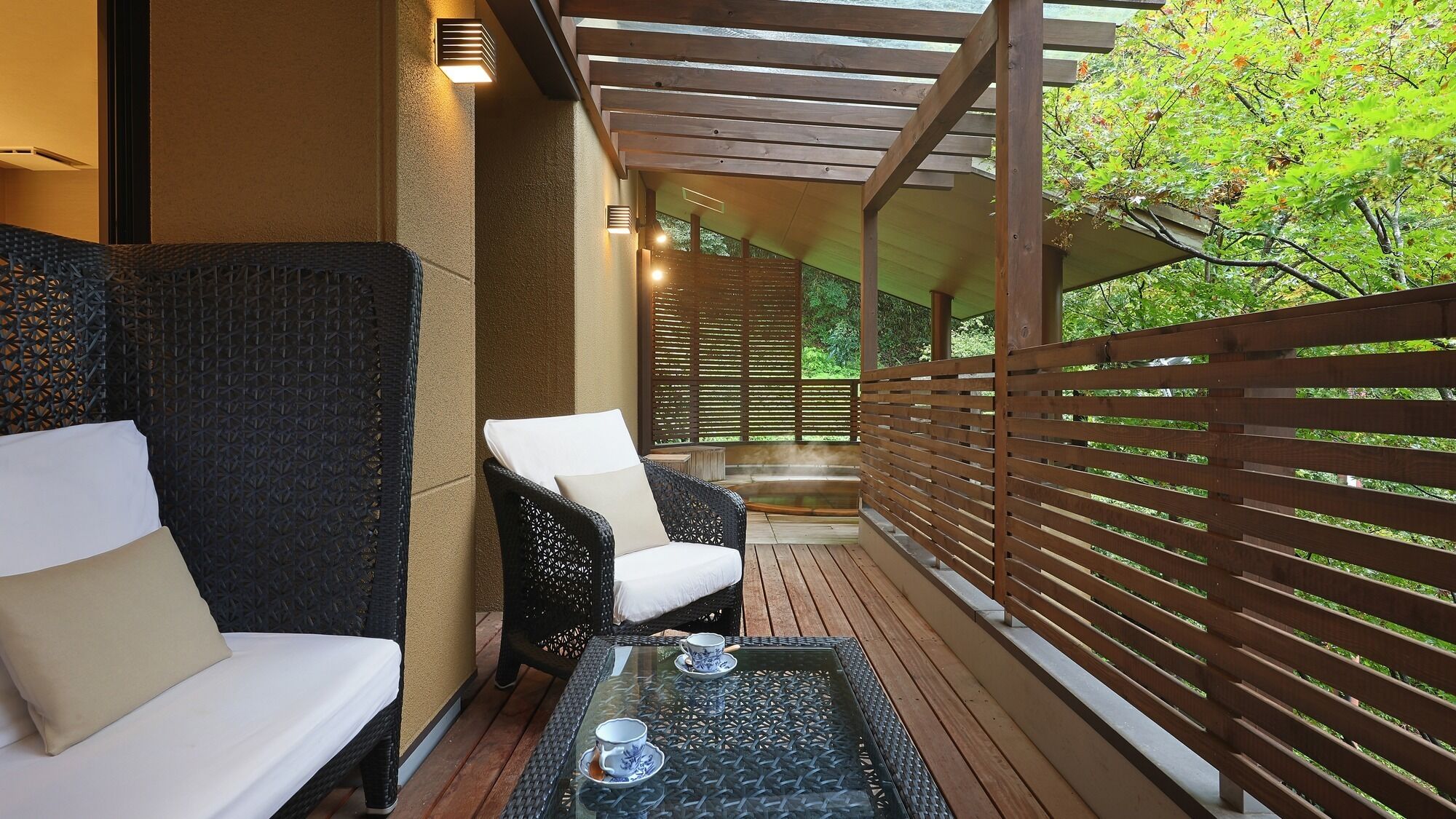 A covered outdoor seating area with wicker furniture and a glass table, surrounded by wooden railings and greenery.