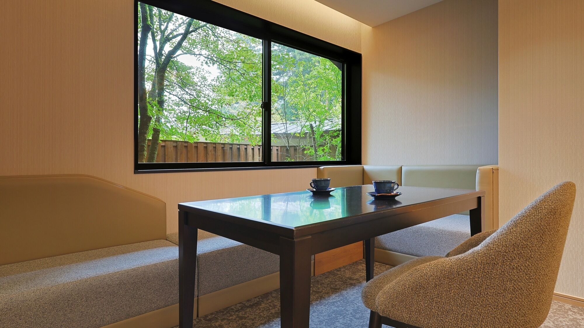 A room with a glass-top table, two chairs, a bench, and a large window with a view of trees and a wooden fence.