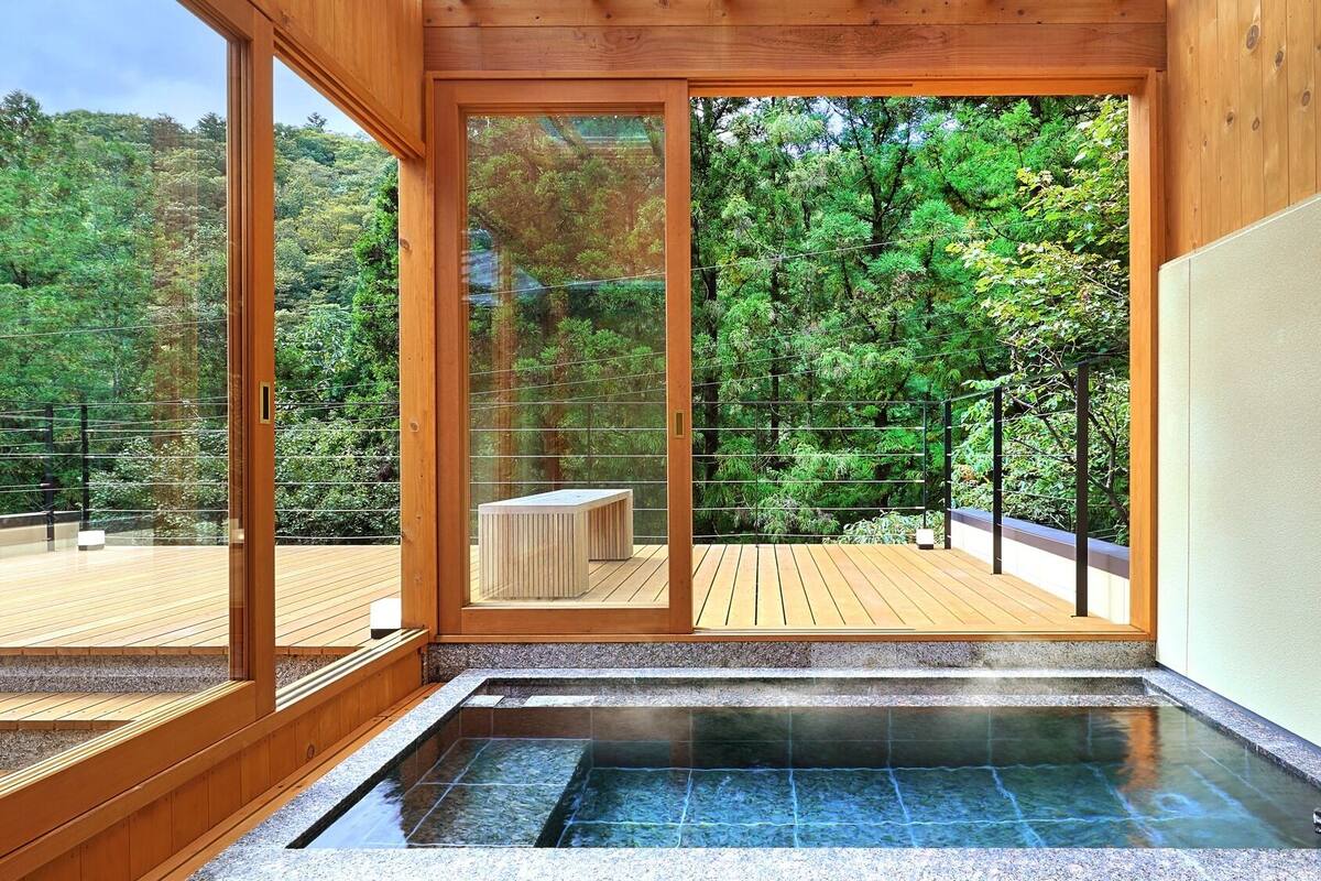 Ein Wellnessbereich mit Whirlpool, Holzterrasse und Blick auf eine bewaldete Landschaft durch große Glaswände.