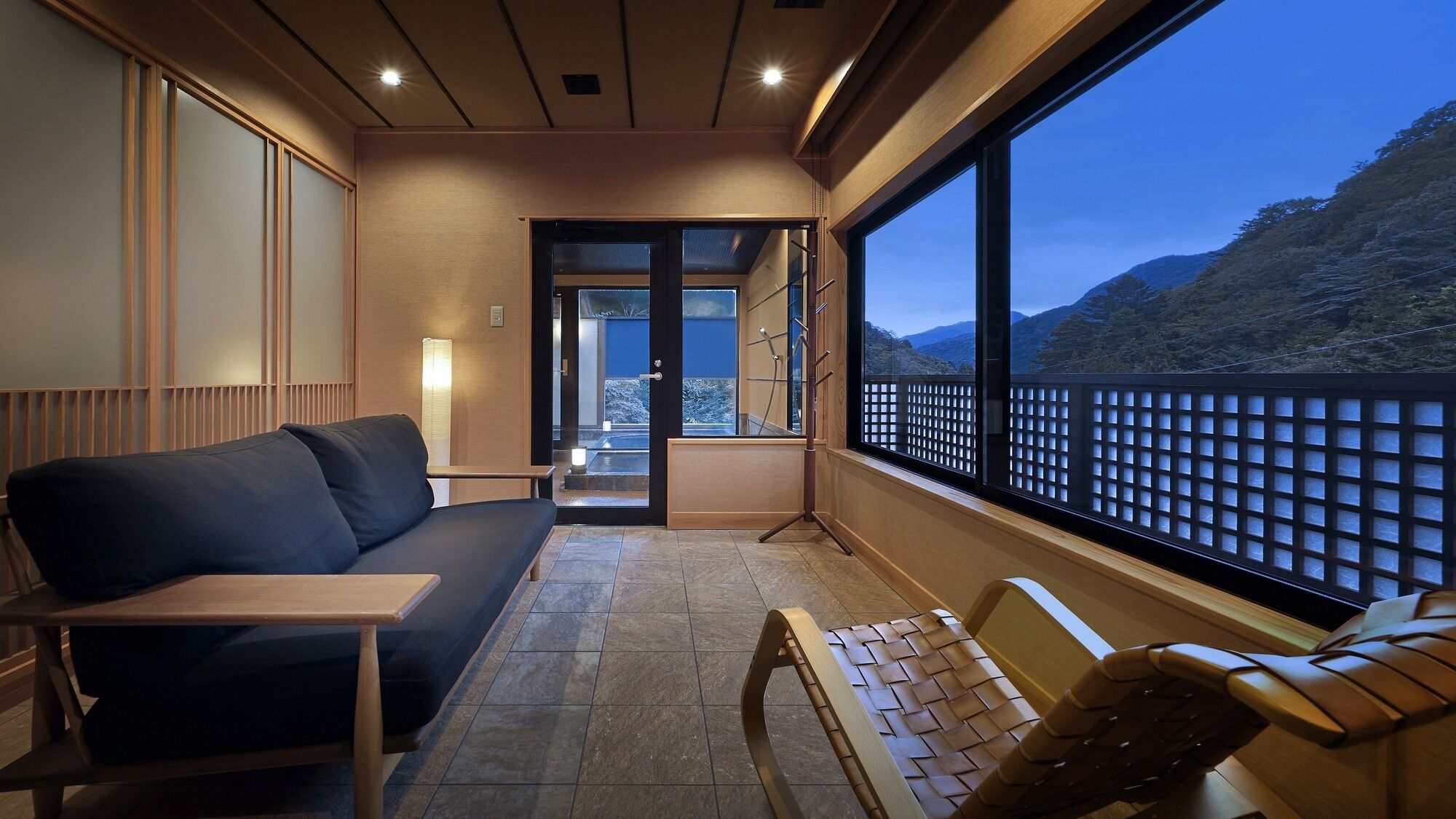 A balcony with a sofa, a small table, and a chair. There is a view of a mountainous landscape through large windows.
