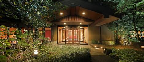 A traditional Japanese-style building with a wooden entrance gate, surrounded by lush greenery and illuminated by soft lighting.