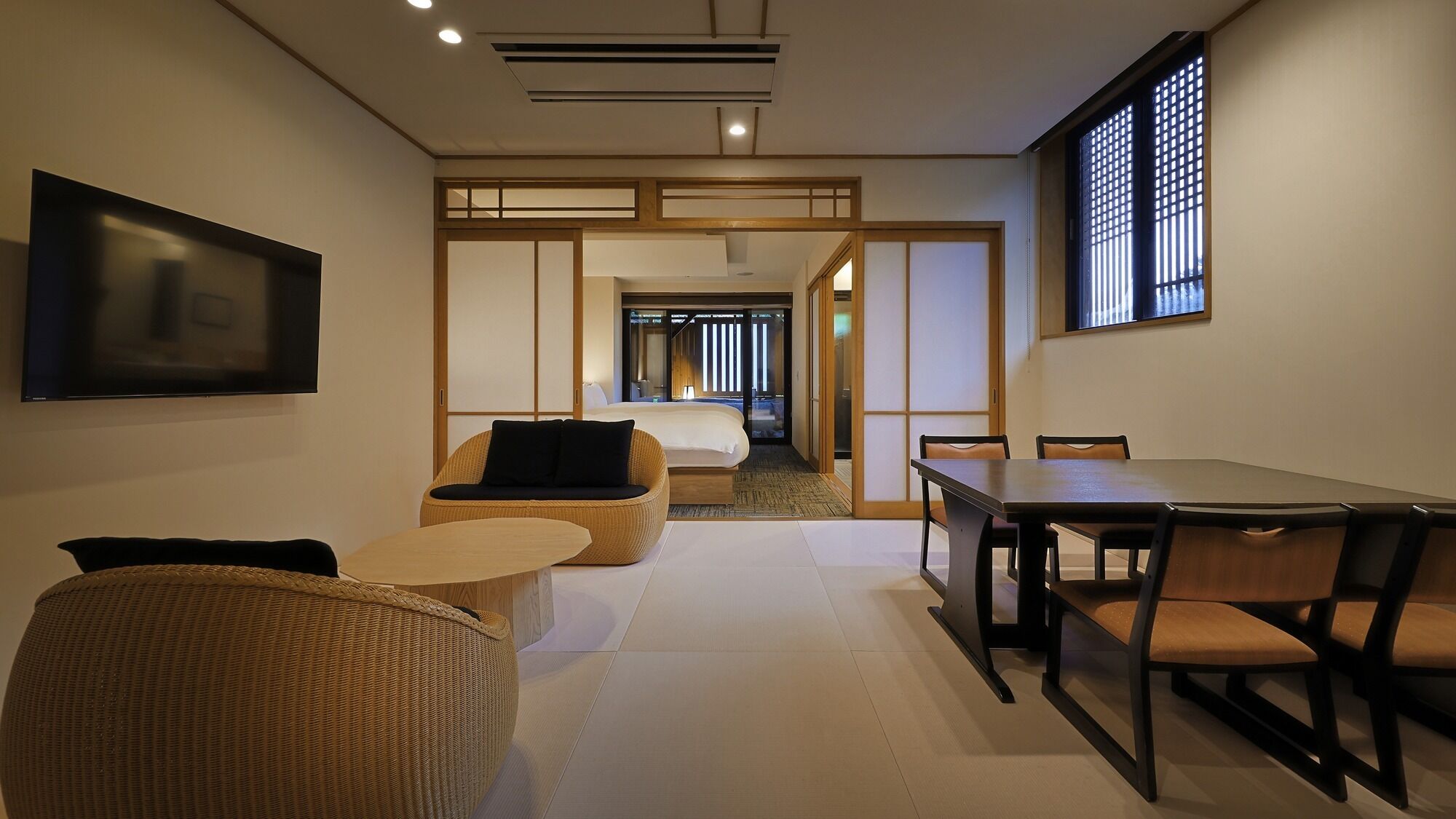 A hotel room with a bed, a dining table, chairs, a TV, and a sliding door leading to another room.