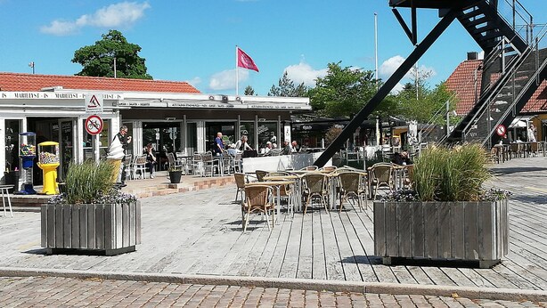 Un café al aire libre con mesas y sillas, una máquina expendedora amarilla e un edificio con una bandera roja.