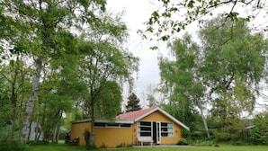 Una pequeña casa amarilla con techo rojo, rodeada de árboles y césped.