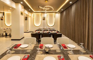 Un espace de restauration moderne avec une longue table dressée pour un repas, agrémenté d’un luminaire central et offrant une vue sur un mur recouvert de rideaux.