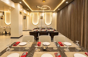 A modern dining area with a long table set for a meal, featuring a central light fixture and a view of a curtain-covered wall.