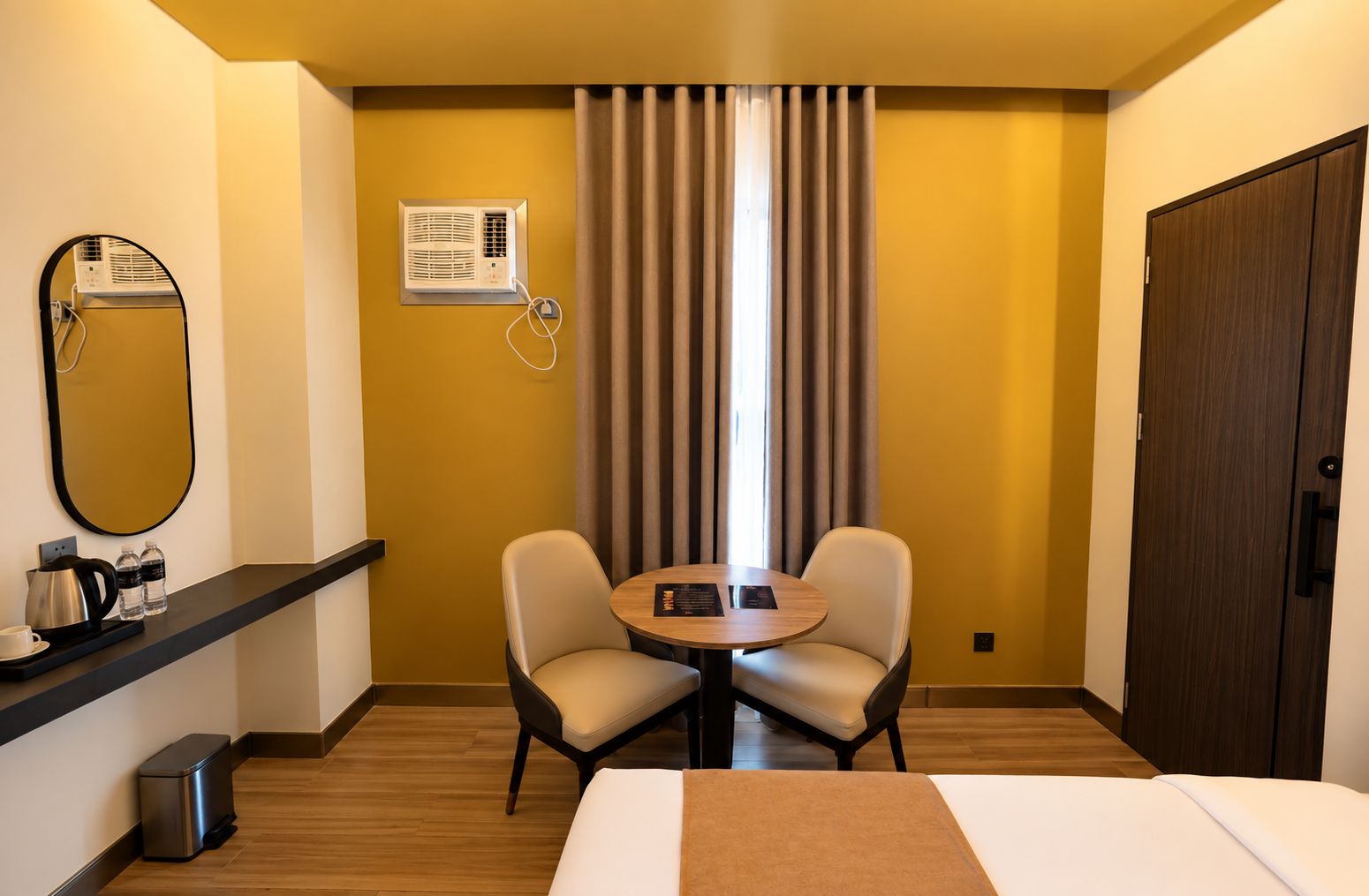 A hotel room with a bed, two chairs, a small table, a mirror, a coffee maker, and a trash bin.
