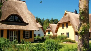 A row of traditional thatched-roof cottages with white walls and green shutters, surrounded by lush greenery. - Küstenwaldvilla in Dierhagen (Dierhagen)