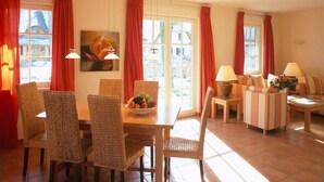 A dining area with wicker chairs, a wooden table, and a fruit bowl. There are two windows with red curtains, and a painting on the wall. - Küstenwaldvilla in Dierhagen (Dierhagen)