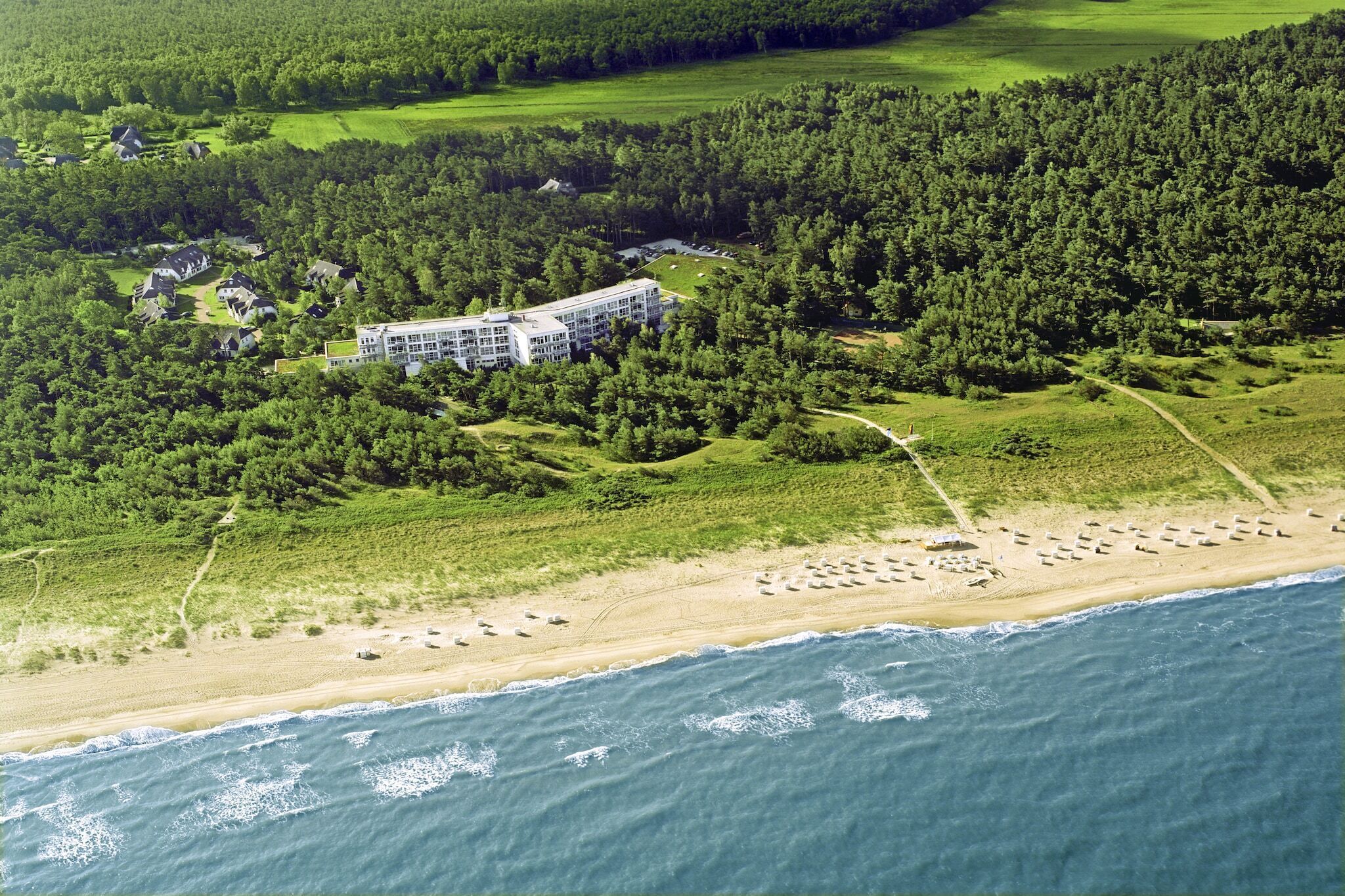 Vista aérea de un complejo turístico costero con un edificio grande rodeado de bosque denso, una playa de arena y una vista despejada del océano.