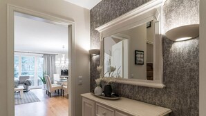 A hallway with a large mirror, a white console table with decorative items, and a view into a dining area with a chandelier.