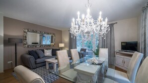A modern living room with a glass dining table, a chandelier, a sofa, and a television.