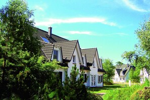 A row of white houses with dark roofs and multiple chimneys, surrounded by lush greenery and trees.