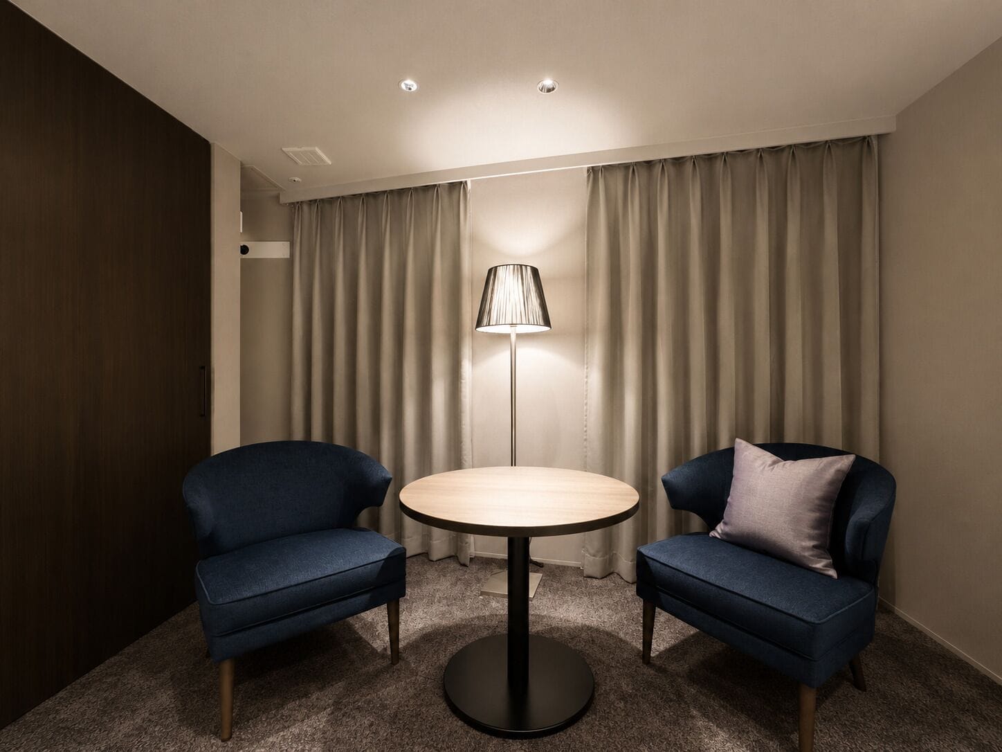 Una habitación con dos sillones azules, una mesa redonda, una lámpara de pie y cortinas color beige.