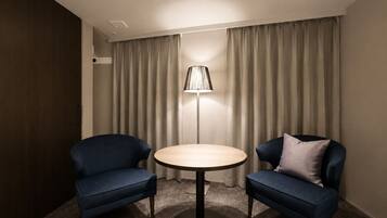 Una habitación con dos sillones azules, una mesa redonda, una lámpara de pie y cortinas color beige.