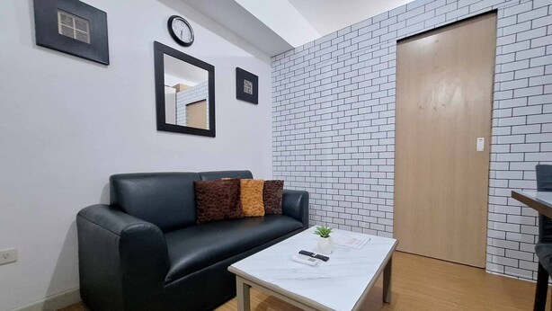 Une pièce avec un canapé en cuir noir, une table basse blanche, une horloge, un miroir et une porte donnant sur un mur en briques.