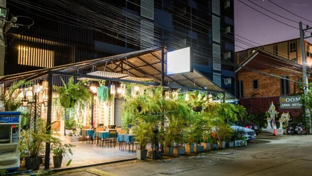 Restaurante com mesas ao ar livre, rodeado por vasos de plantas, e um letreiro do hotel 'ROOM' ao fundo.
