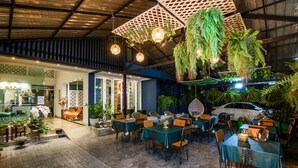Um restaurante interno com mesas e cadeiras, plantas penduradas e um carro estacionado na rua.