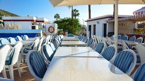 Zona de comedor junto a la piscina, con mesas blancas y sillas a rayas azules, rodeada de palmeras y un cielo despejado.