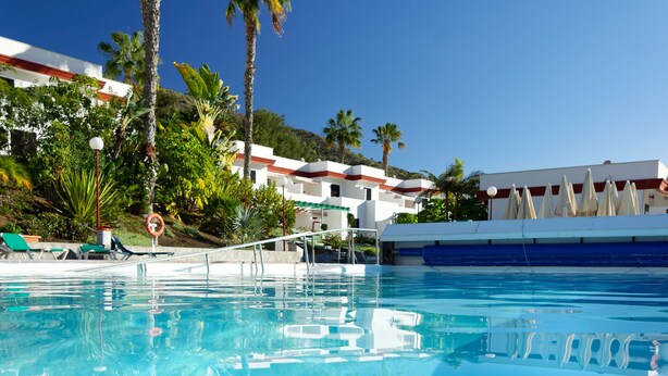 A swimming pool with lounge chairs and umbrellas, surrounded by palm trees and a building with a red roof. - VALLE MARINA APARTAMENTOS (Arguineguin)