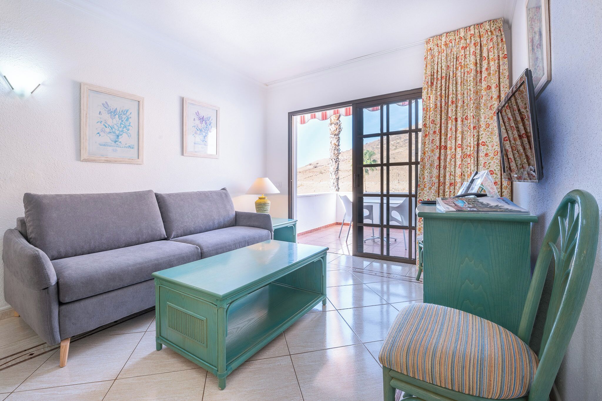 A living room with a grey sofa, a green coffee table, and a striped chair. There is a window with a view of a desert landscape.
