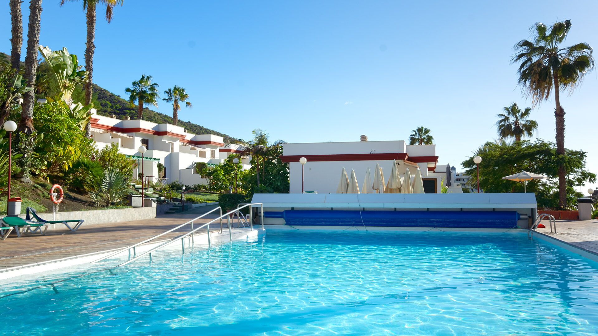 Una piscina con cubierta azul y blanca, rodeada de palmeras y un edificio con techo rojo.