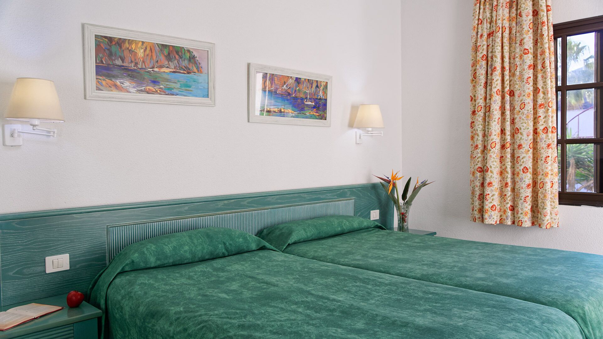 Habitación de hotel con una cama tapizada de verde, dos lámparas fijadas a la pared, dos cuadros enmarcados, una cortina con motivos florales y una mesita de noche con un libro y una manzana.