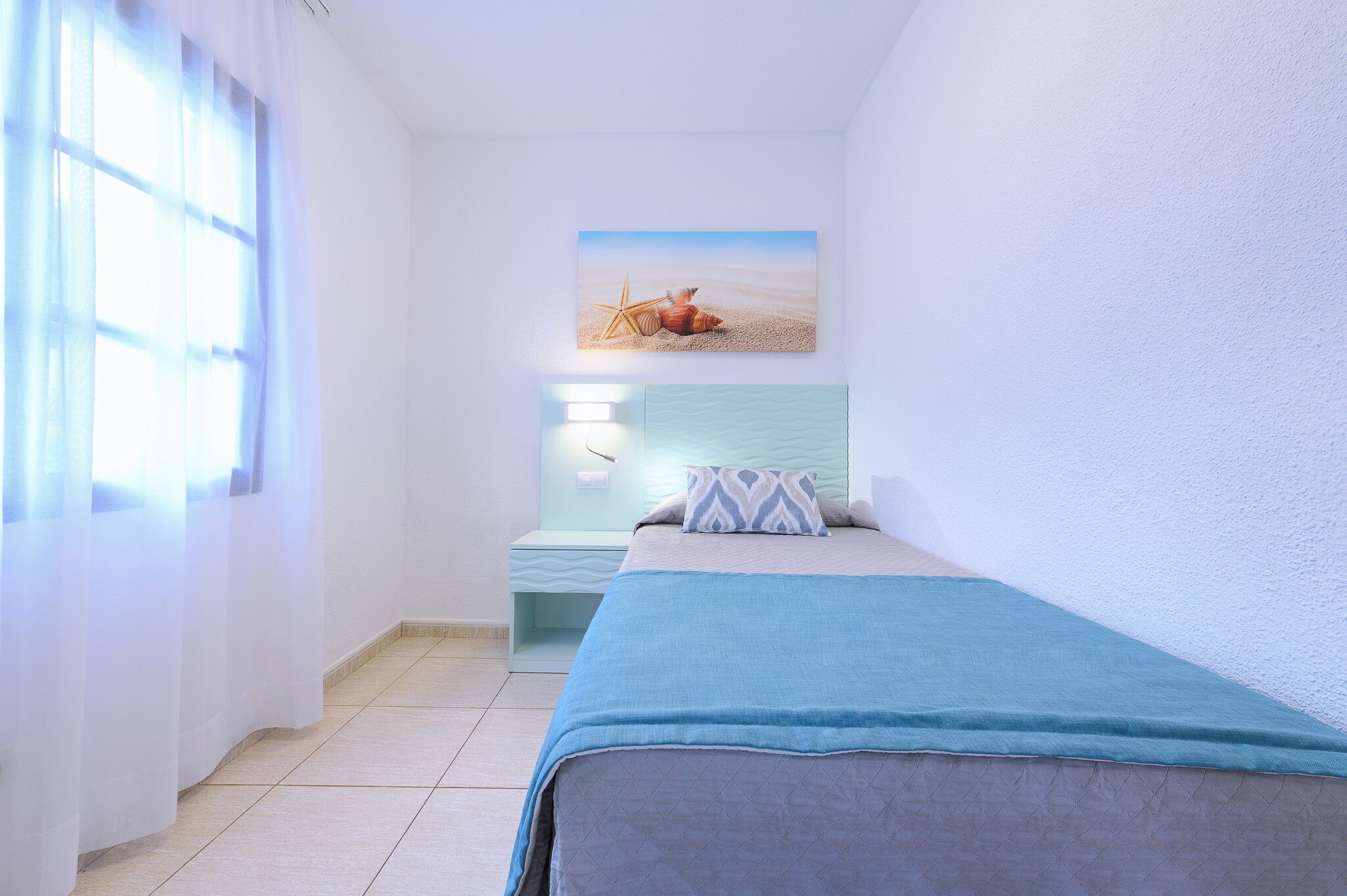 A bedroom with a bed, a nightstand, a wall-mounted lamp, and a beach-themed painting.