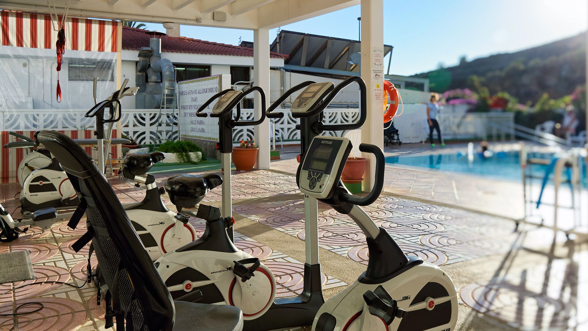 An outdoor gym area with stationary bikes and a rowing machine, near a pool and a covered seating area.