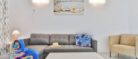 A modern living room with a grey sofa, a white coffee table, and a beige armchair. There is a colorful blanket draped over the sofa, a hat, and a decorative pillow. A painting hangs on the wall above the sofa.