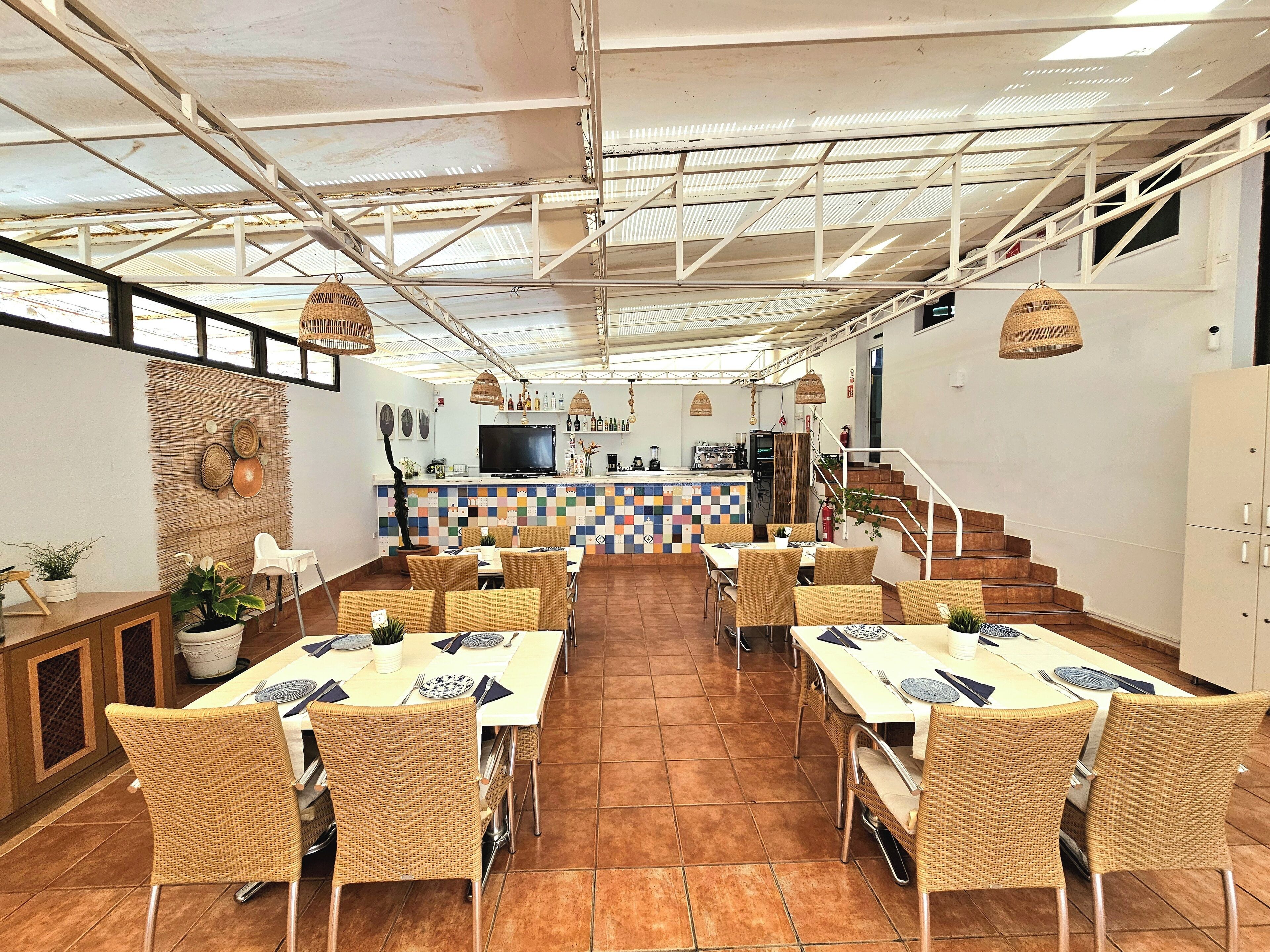 Un restaurante con techo alto, vigas a la vista y una zona de bar central. Las mesas están dispuestas con sillas de mimbre y un suelo de baldosas.