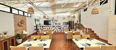 Un restaurante con techo alto, vigas a la vista y una zona de bar central. Las mesas están dispuestas con sillas de mimbre y un suelo de baldosas.