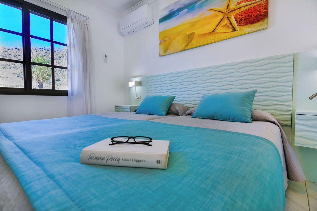 Un dormitorio con cama, ventana con vista a paisaje desértico, pintura en la pared y mesita de noche con lámpara.