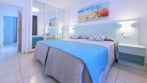 A bedroom with a bed, a nightstand, a mirror, and a painting of a beach scene. - VALLE MARINA APARTAMENTOS (Arguineguin)
