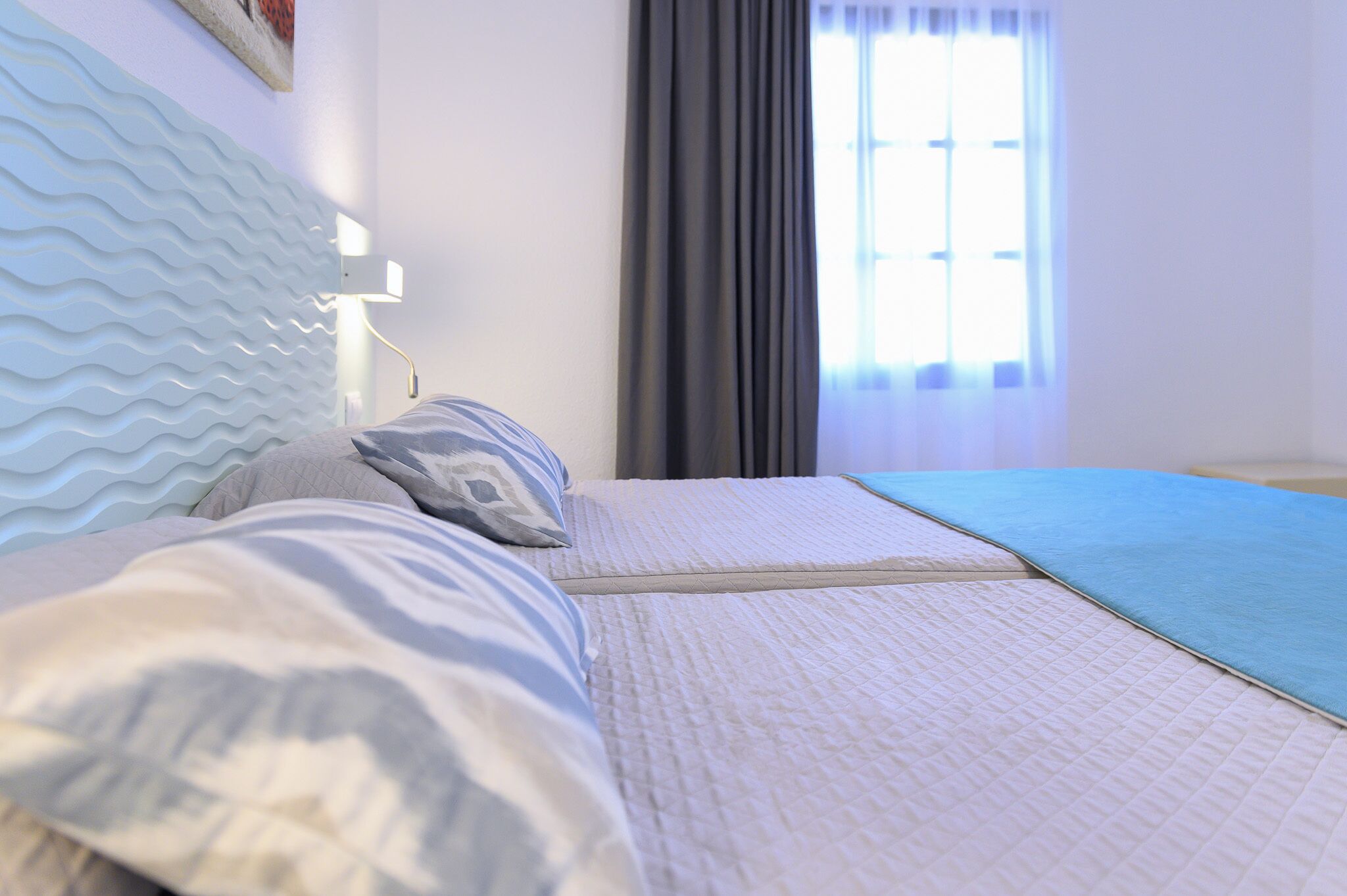 Ein Hotelzimmer mit einem Bett, einem Wellenmuster-Kopfstück, einer Nachttischlampe, einem Fenster mit Vorhängen und einer blauen Decke auf dem Bett.