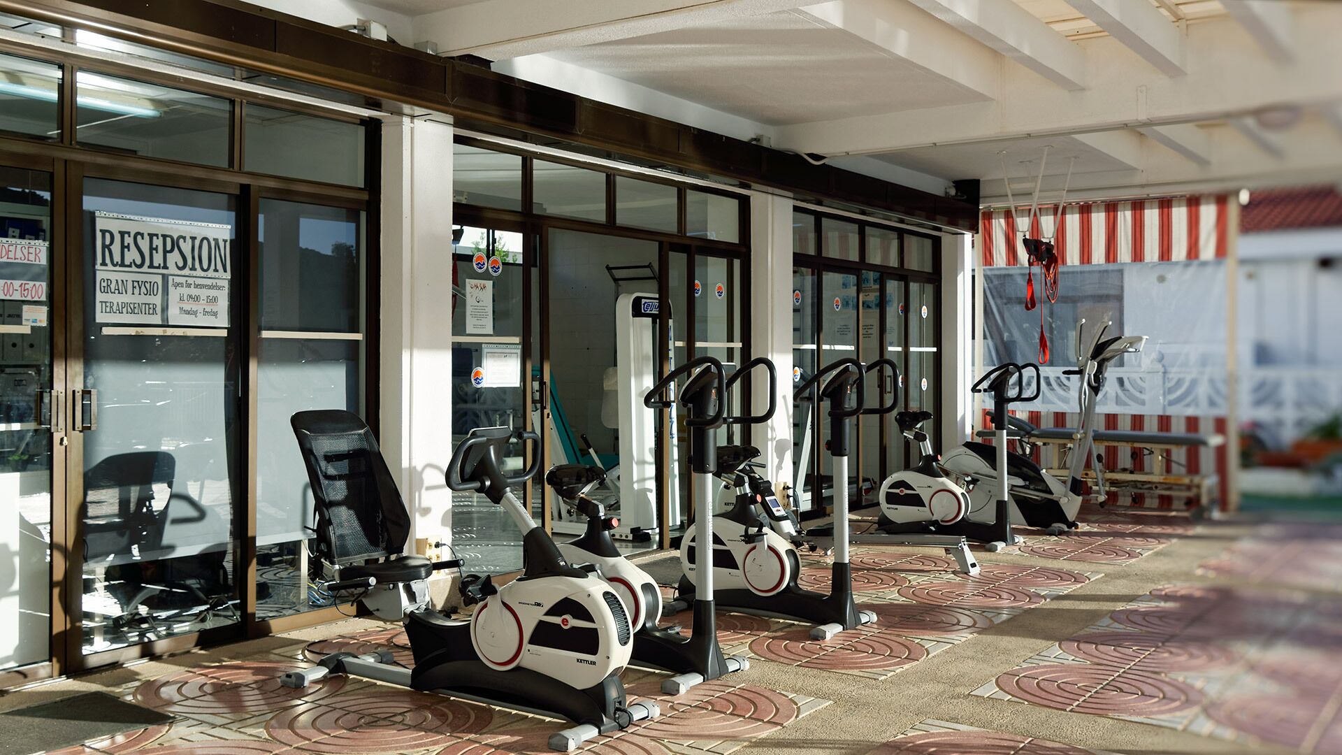 A gym with various exercise machines, a reception area, and a patterned floor.
