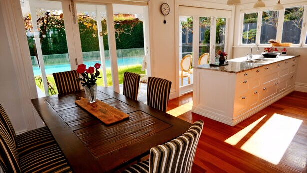 Un comedor con mesa de madera, sillas a rayas y un jarrón con flores rojas. Se ve una piscina y vegetación al exterior.